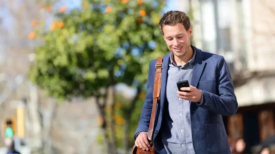 Young professional walking while outside. He's looking at his cell phone and carrying a work bag.