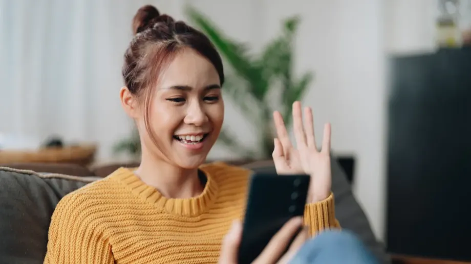 Young woman wearing a gold sweater and holding her phone. She's waving at the phone while talking to her community.